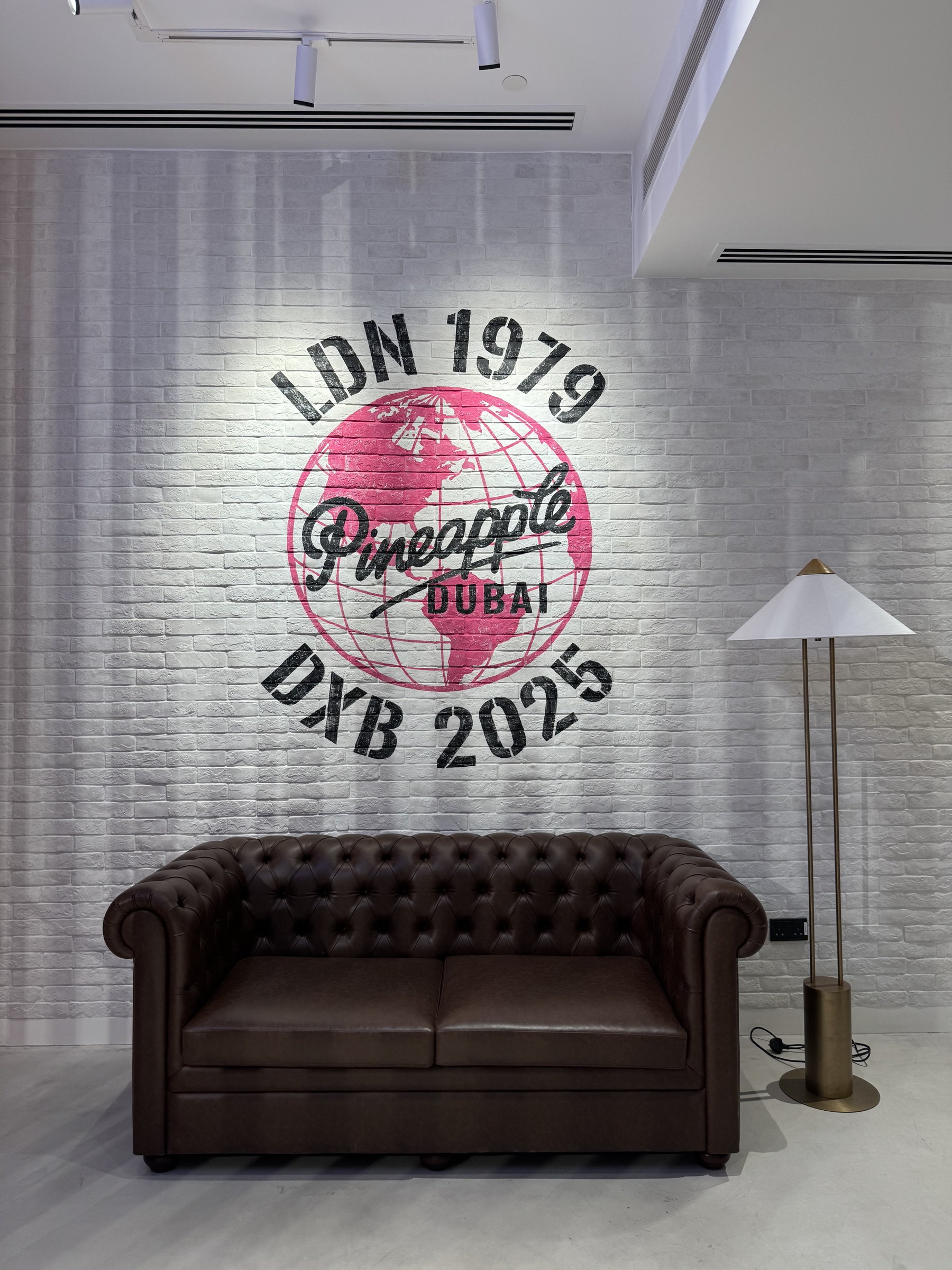 Pineapple 'LDN 1979 · DXB 2025' crest painted on a white wall above a leather sofa